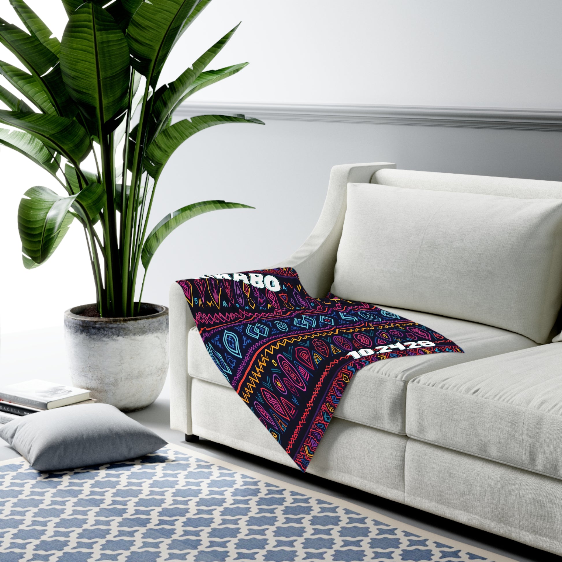A white sofa with a colorful patterned swaddle blanket draped over one arm sits next to a large potted plant. A blue-and-white geometric rug covers the floor, and a gray floor cushion rests nearby in this modern, African-inspired living room.