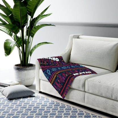 A white sofa with a colorful patterned swaddle blanket draped over one arm sits next to a large potted plant. A blue-and-white geometric rug covers the floor, and a gray floor cushion rests nearby in this modern, African-inspired living room.
