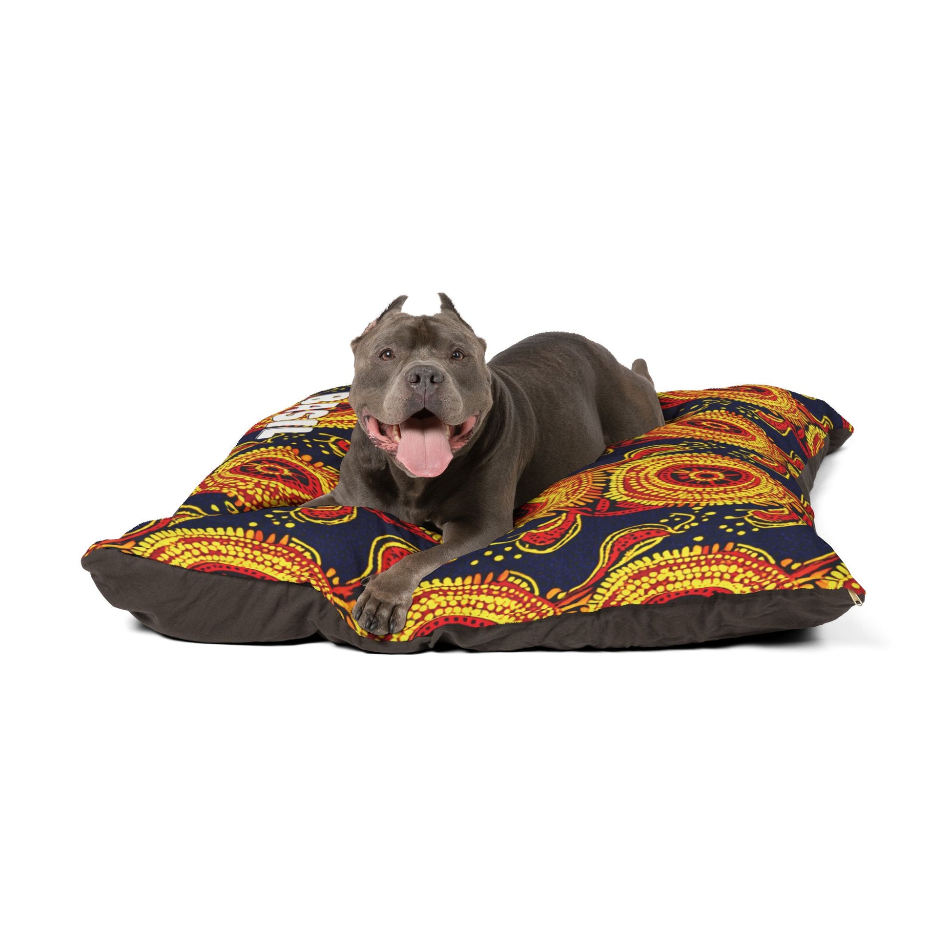 A gray dog with cropped ears lies on a colorful, African-inspired patterned cushion. The dog looks forward with its mouth open and tongue out, appearing happy and relaxed against vibrant red, yellow, and orange circular designs on navy.
