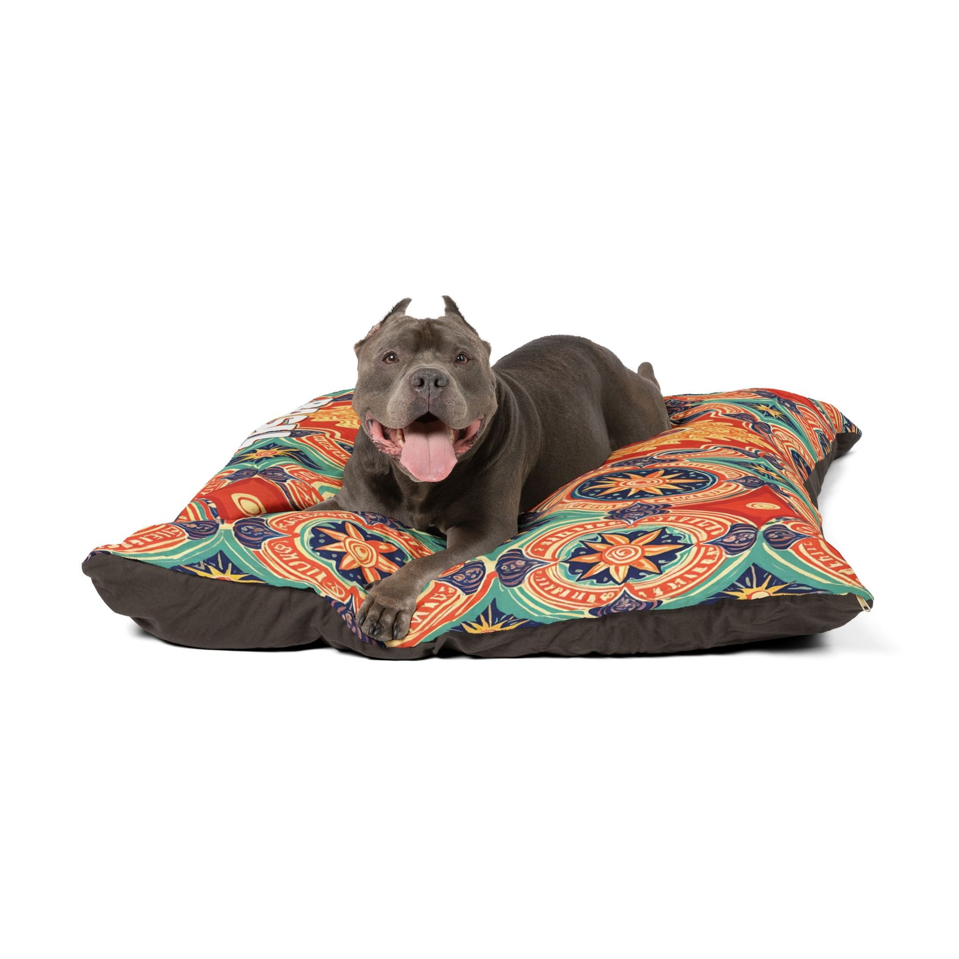 A gray dog with cropped ears lies on a stylish pet bed, facing forward with its mouth open and tongue out, appearing happy and relaxed. The cozy dog bed features bright geometric and floral designs in red, orange, blue, and green.