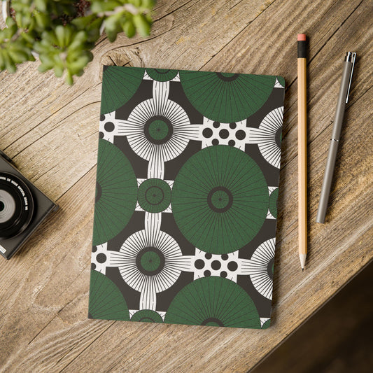 An African-inspired journal with a bold geometric pattern of green circles, white shapes, and black dots lies on a rustic wooden desk, perfect for daily journaling or as a travel diary. Nearby are a pencil, gray pen, camera, and a green potted plant.