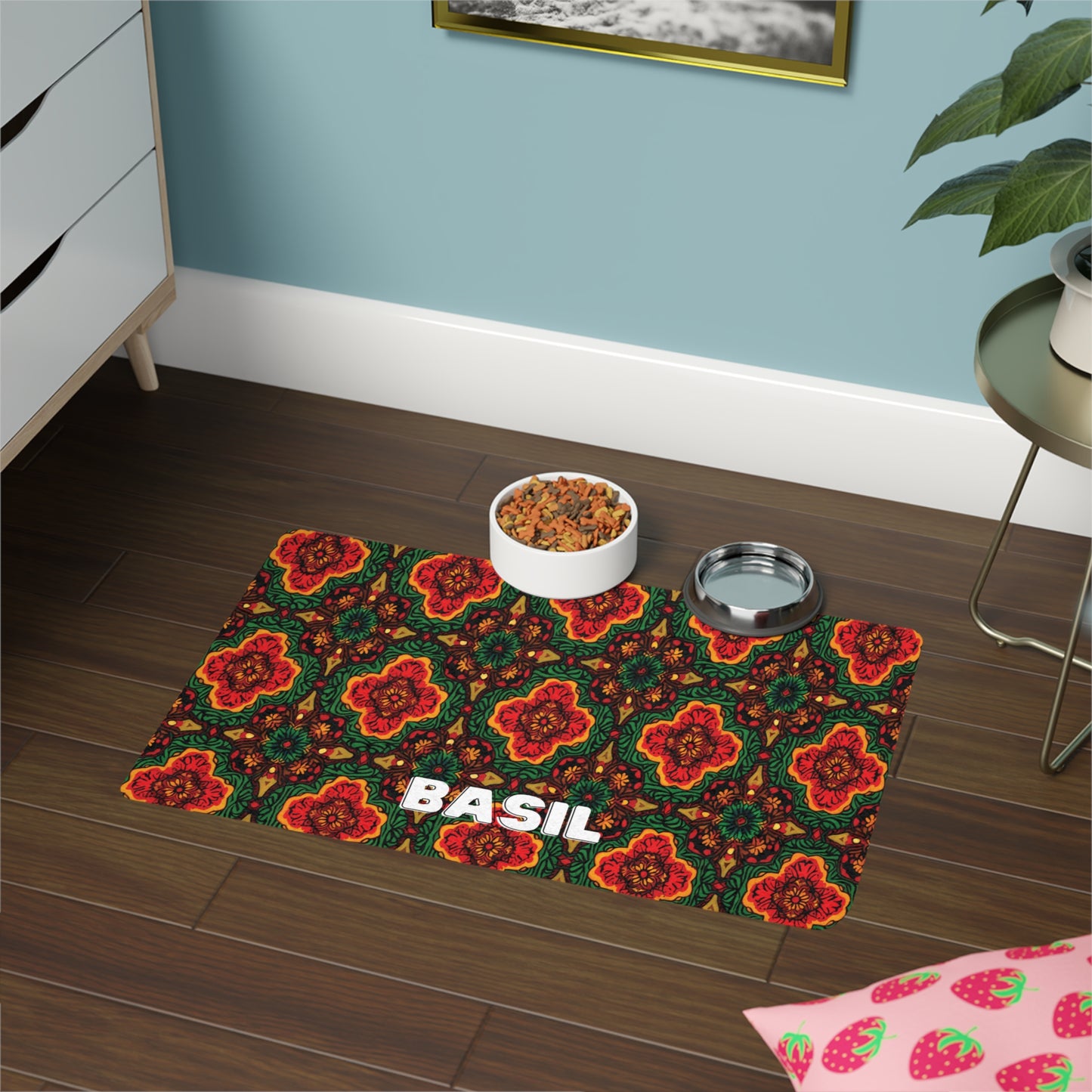 A colorful, African-inspired pet mat with a red, orange, and green floral pattern and the name BASIL in bold white letters sits on a dark wooden floor. On the mat are bowls of pet food and water, with nearby furniture, a plant, and a pink strawberry-patterned cloth.
