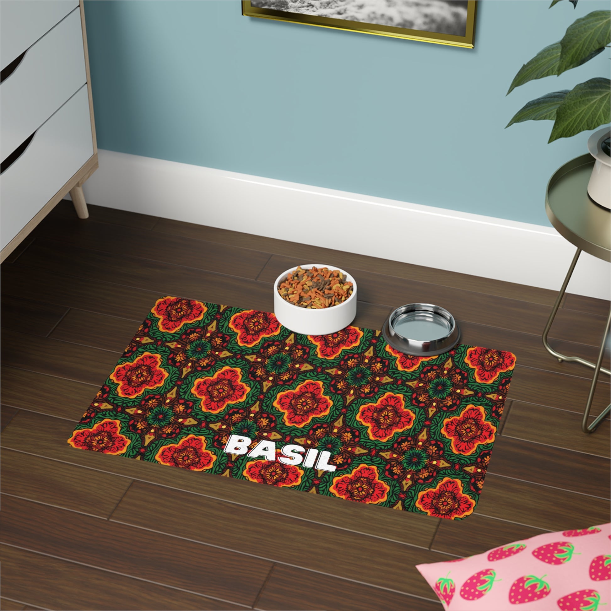 A colorful, African-inspired pet mat with a red, orange, and green floral pattern and the name BASIL in bold white letters sits on a dark wooden floor. On the mat are bowls of pet food and water, with nearby furniture, a plant, and a pink strawberry-patterned cloth.