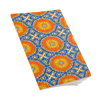 A creative journal with a vibrant cover featuring a symmetrical pattern of orange and yellow sunburst shapes, blue geometric details, and white star-like accents. The African-inspired journal is closed and photographed at an angle against a white background.