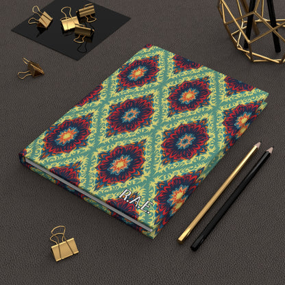 A vibrant African-inspired personalized journal with a colorful geometric floral pattern lies on a textured brown surface. Nearby are gold binder clips, black and gold pencils, and a gold wire decor piece. The notebook’s cover reads PLACE.