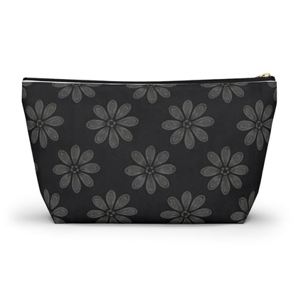 A black cosmetic travel bag featuring a repeating pattern of hand-drawn, grey six-petal flowers with subtle shading. This T-bottom pouch has a wide base, tapers toward the top, and closes with a gold-colored zipper on its solid black background.