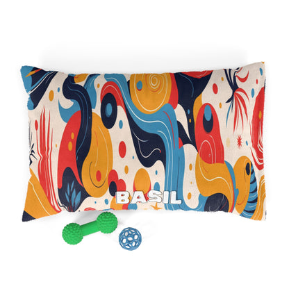 A rectangular pillow with BASIL written in white letters at the bottom center, featuring an African-inspired abstract, multicolored pattern in blue, red, yellow, and beige. Two dog toys—a green bone and a blue ball—are placed in front of the pillow.