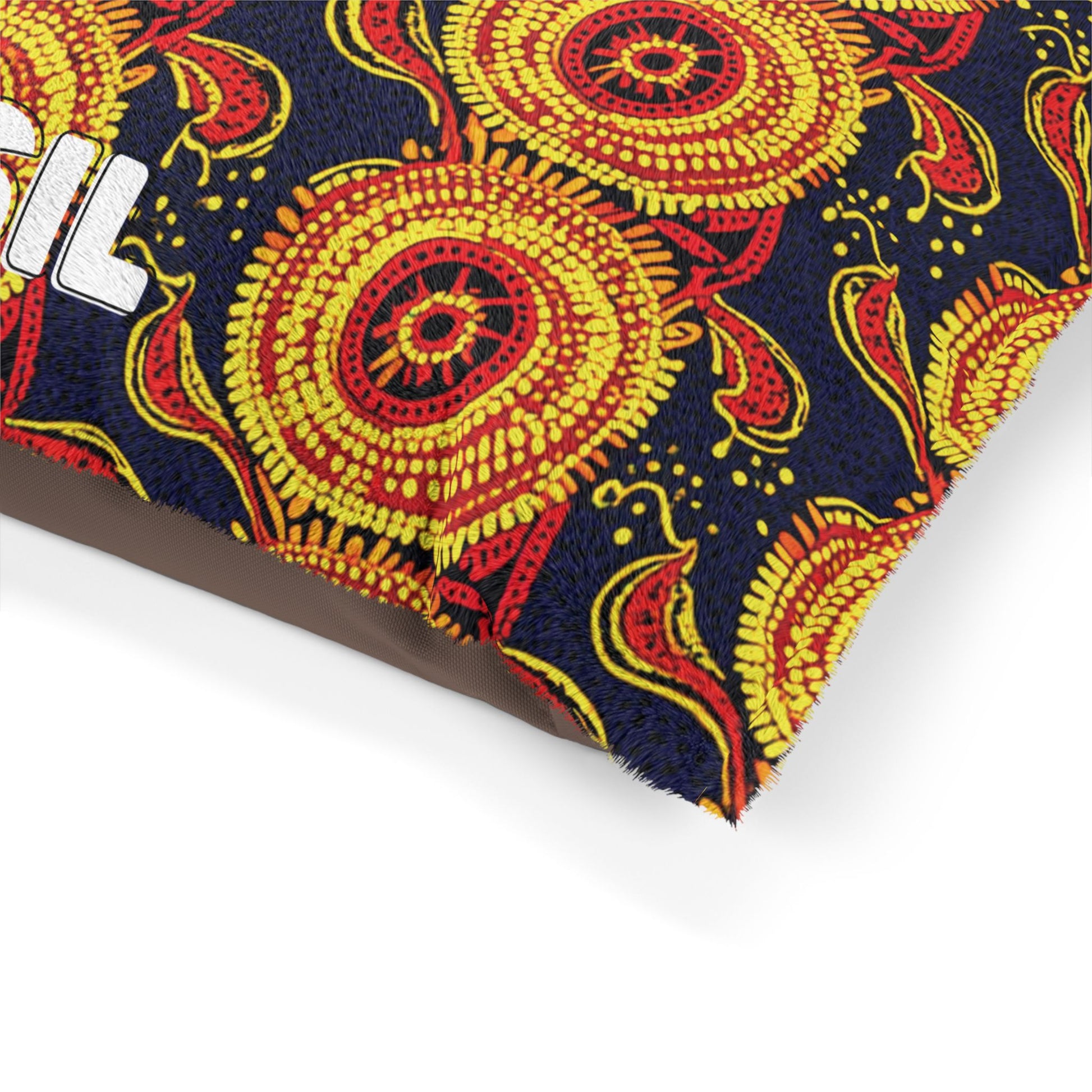 A close-up of a colorful, African-inspired fabric with a bold, embroidered pattern of yellow and red circular floral designs on a dark blue background. Part of a white embroidered word is visible on the left edge, covering what appears to be a cushion or pillow.