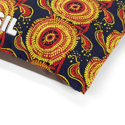 A close-up of a colorful, African-inspired fabric with a bold, embroidered pattern of yellow and red circular floral designs on a dark blue background. Part of a white embroidered word is visible on the left edge, covering what appears to be a cushion or pillow.