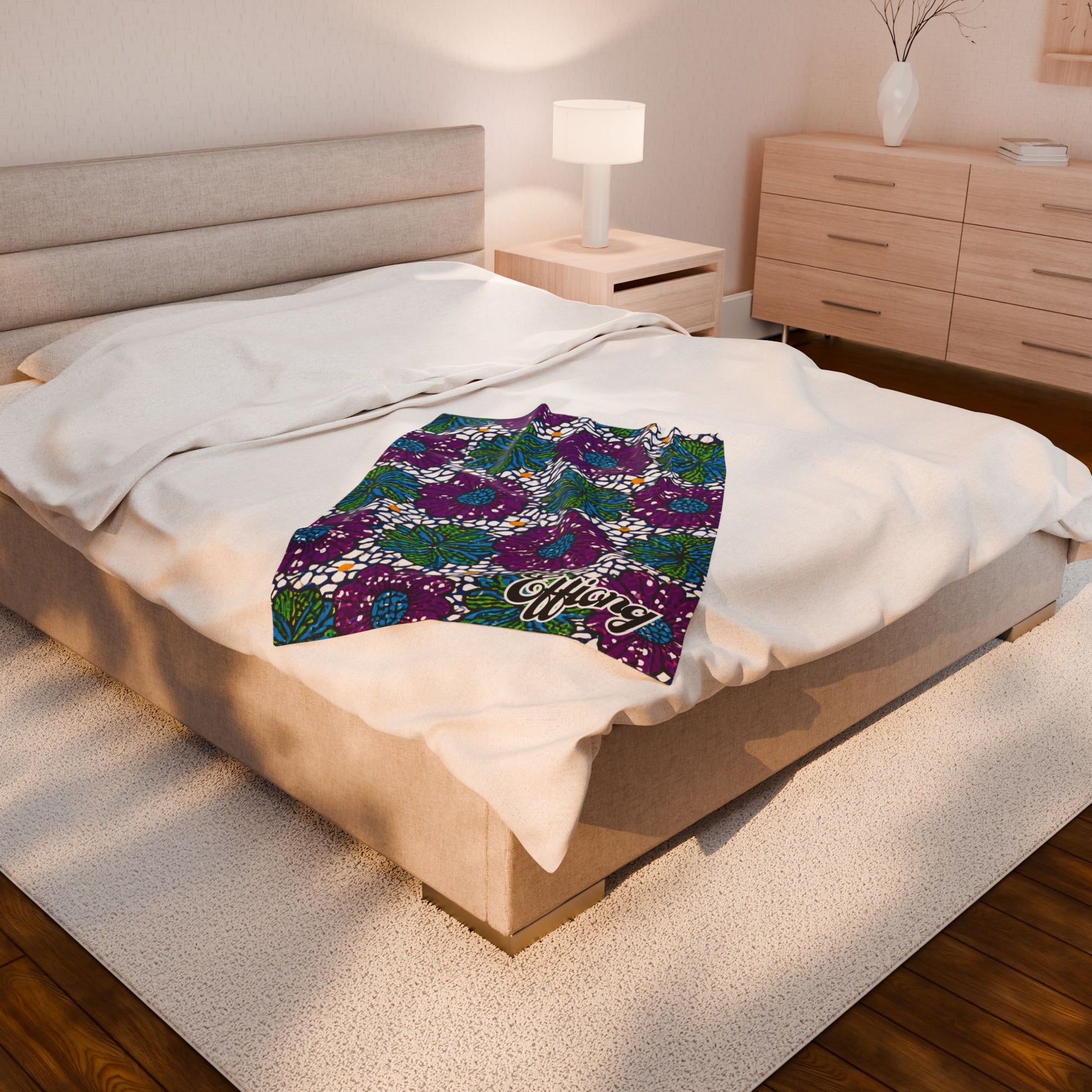 A modern bedroom features a beige bed with white bedding, topped by an African-inspired blanket in green, purple, and white designs. The word “Merry” adds a custom gift touch. Wooden floors, a nightstand with lamp, and a dresser complete the space.