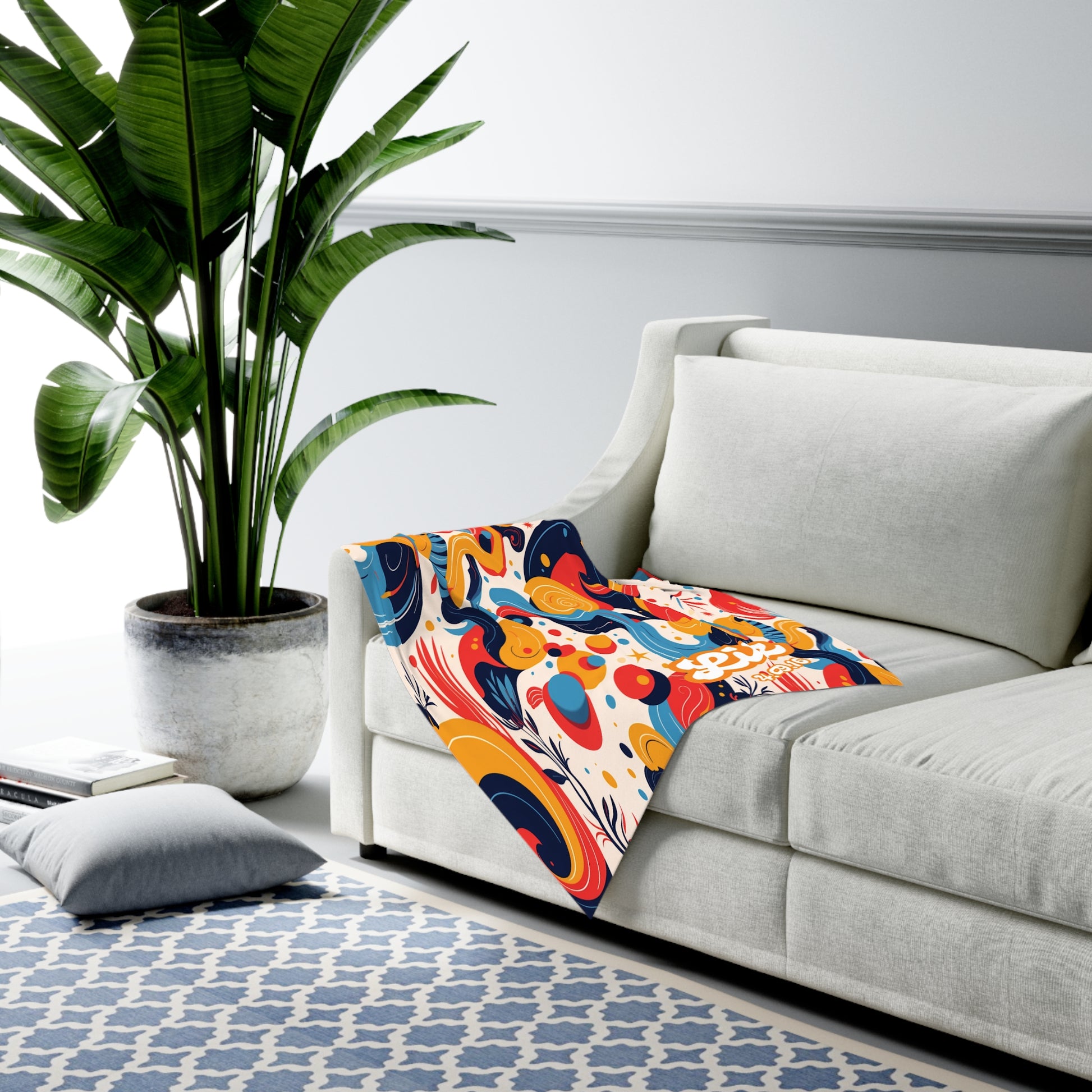 A modern living room with a large green potted plant, a white sofa draped with a colorful abstract-patterned personalized baby blanket, a blue and white geometric rug, a book, and a gray floor cushion. Light walls feature decorative molding.