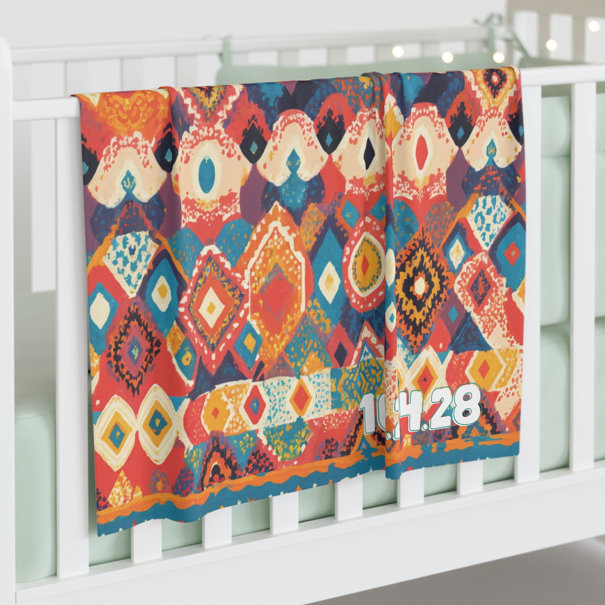 A colorful custom swaddle blanket with an abstract geometric pattern in red, blue, orange, and yellow hangs over a white crib rail. The numbers “1.4.28” are printed in white on the blanket’s corner. The crib mattress is pale mint green.