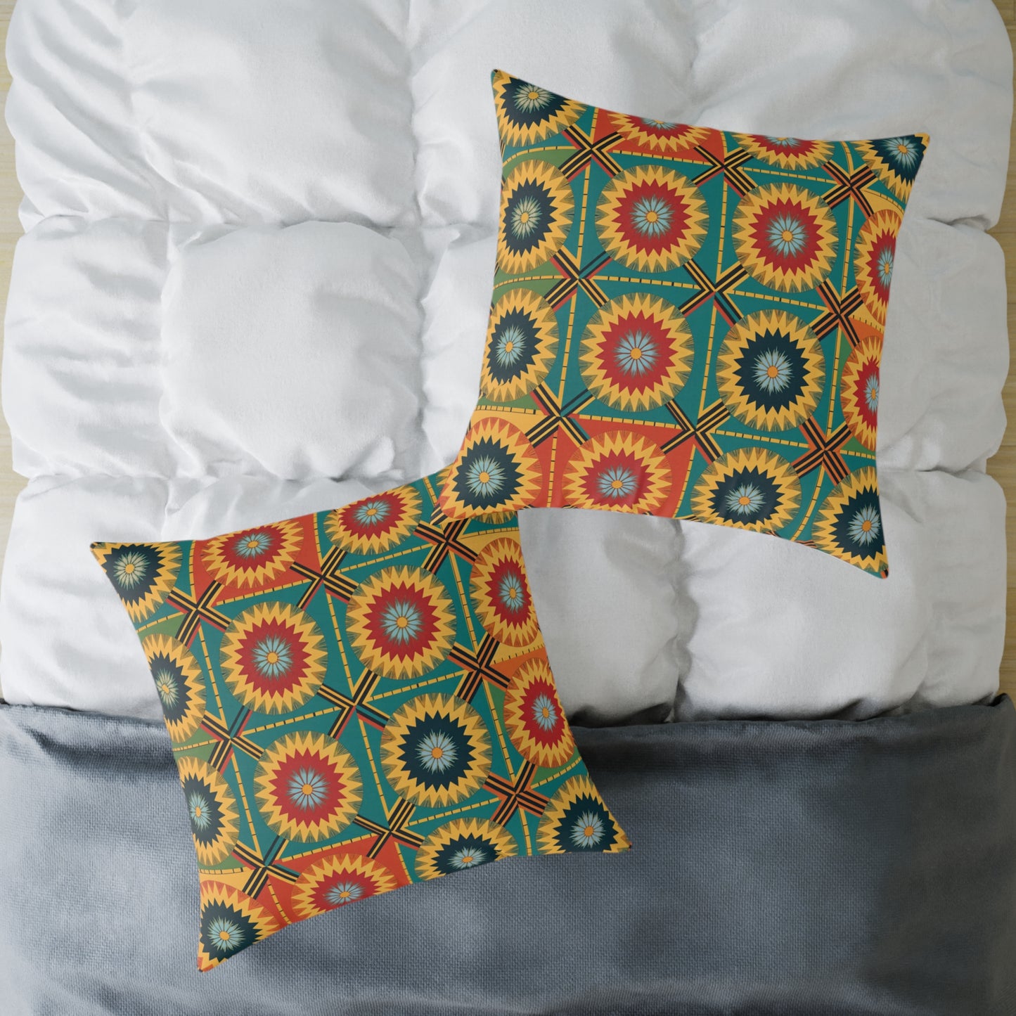 Two square pillows with bold, African-inspired patterns in red, blue, yellow, and green add colorful home decor to a white quilted bed with a gray blanket at the foot. The vibrant throw pillow designs feature concentric starburst and diamond shapes.
