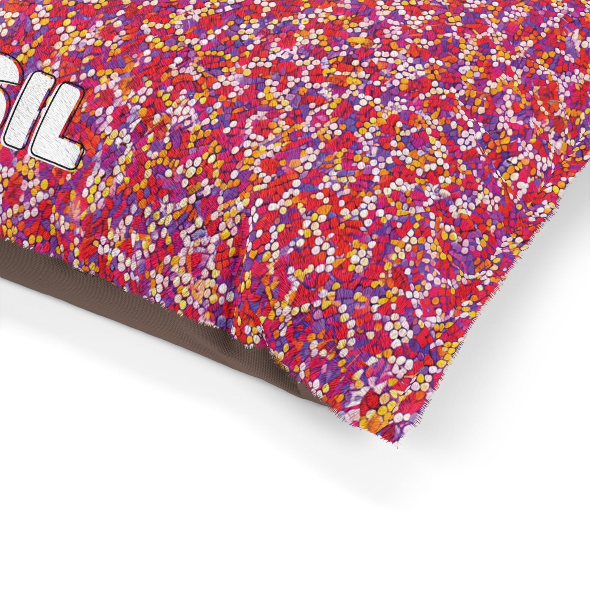 A colorful pillow with durable construction features a dense pattern of tiny red, pink, orange, yellow, and white dots. Partial text SIL in bold white letters appears on the upper left. The pillow sits on a white background with a brown underside.