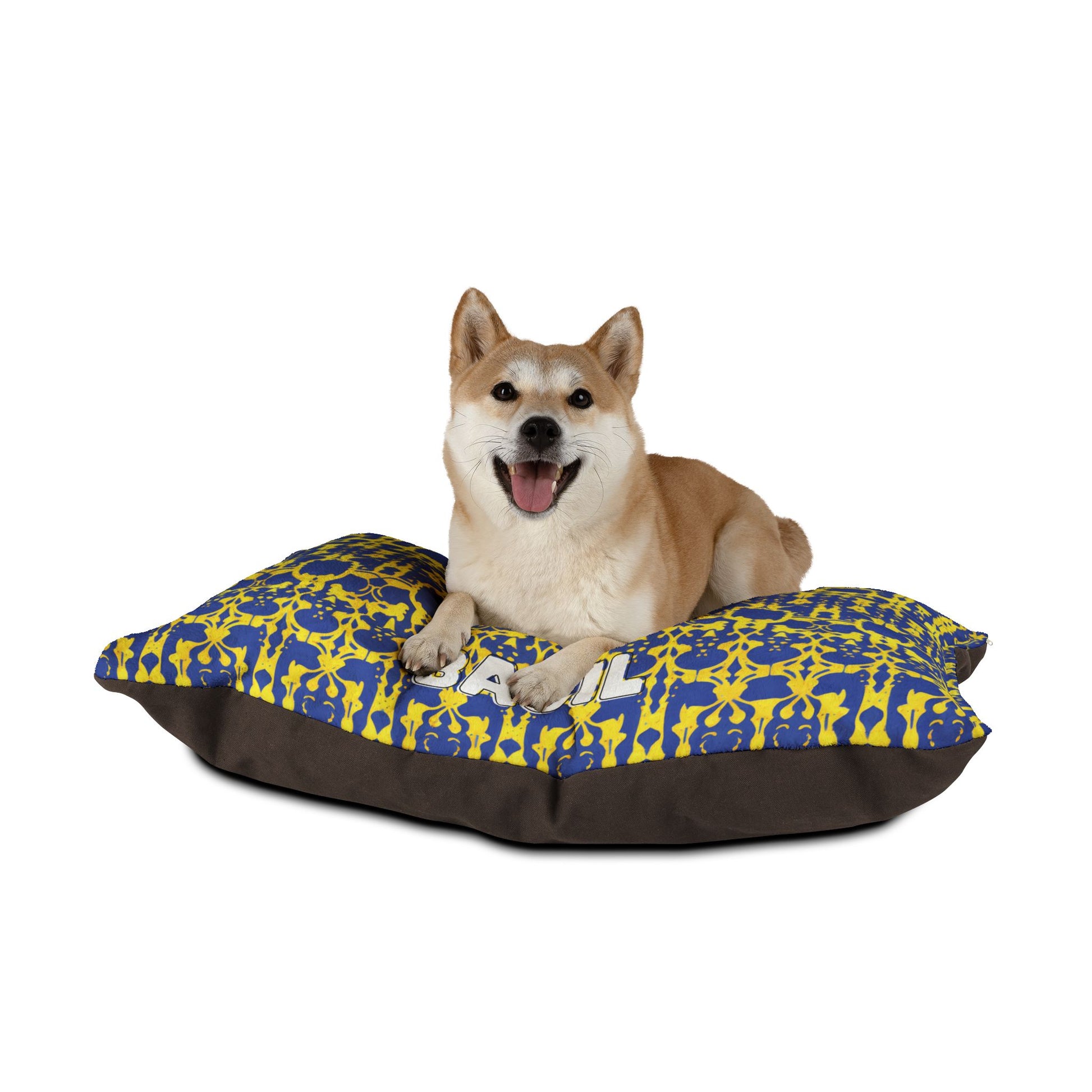 A happy Shiba Inu dog lies on a rectangular cushion with blue and yellow patterns and a solid dark brown base, enjoying luxurious comfort against a plain white background. The dog faces forward with its mouth open and tongue visible.