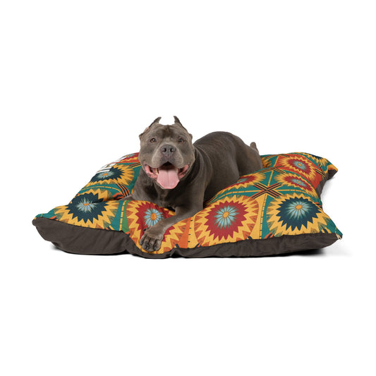 A gray pit bull with cropped ears and a pink tongue lies happily on a stylish pet pillow, featuring red, yellow, green, and blue starburst shapes on a brown background, against a plain white backdrop.