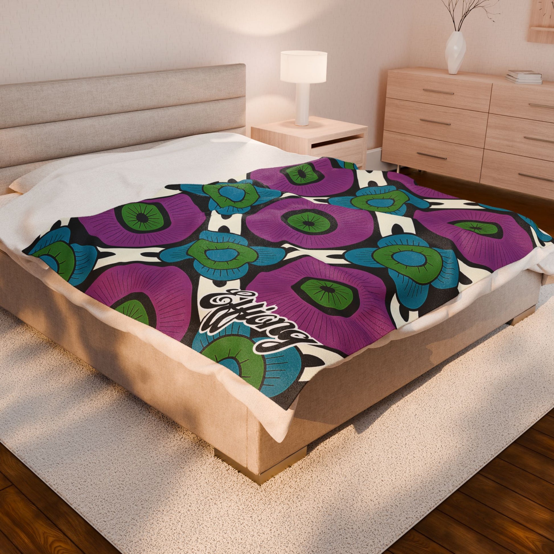 A modern bedroom with a beige bed and matching furniture. A colorful custom name blanket with bold purple, blue, and green floral patterns and the word Happy script covers the bed. A white lamp and minimalist decor add to the cozy, contemporary feel.