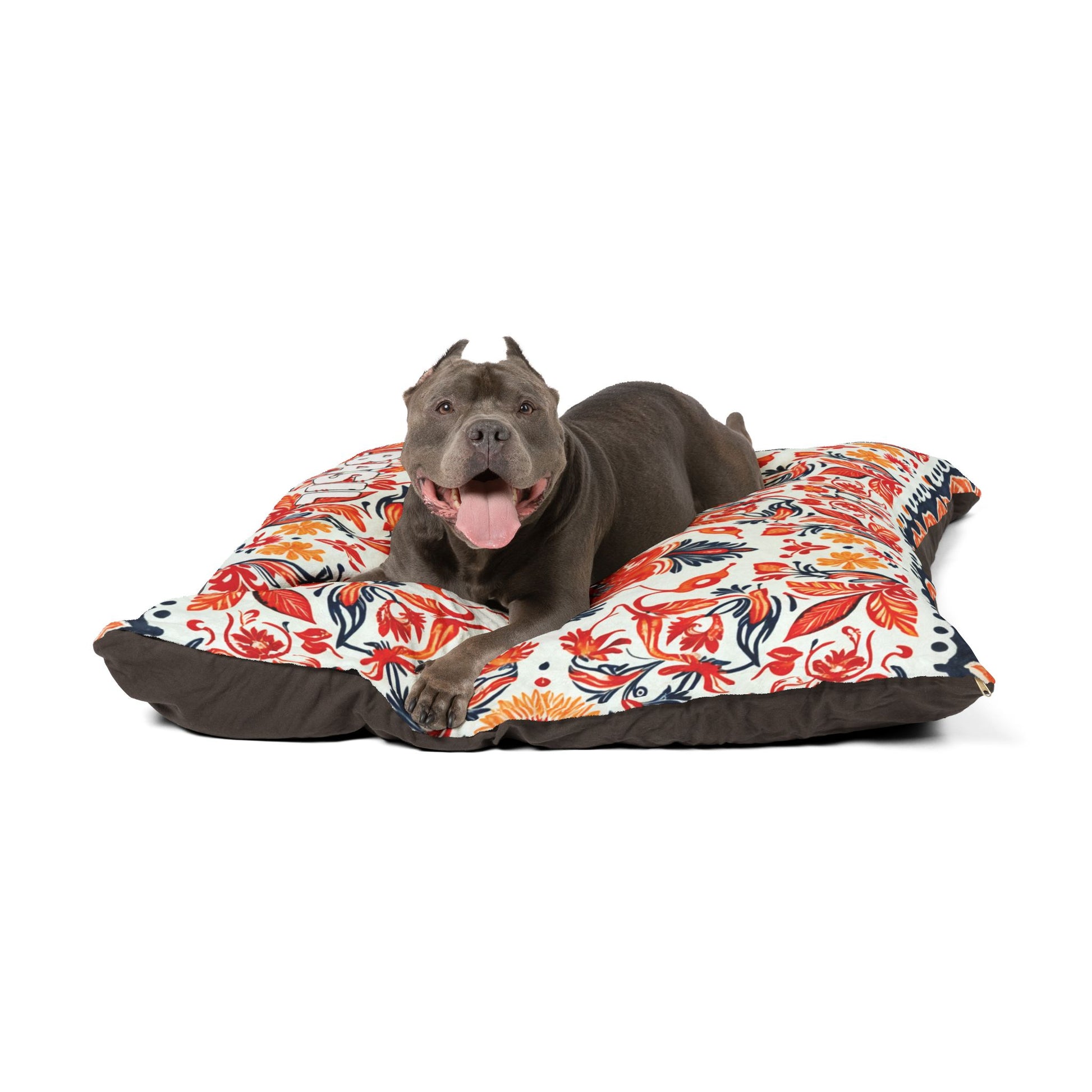 A gray dog with cropped ears and a red collar lies on a large, colorful dog bed with an African-inspired floral pattern. The dog looks happy with its mouth open and tongue out, set against a plain white background.