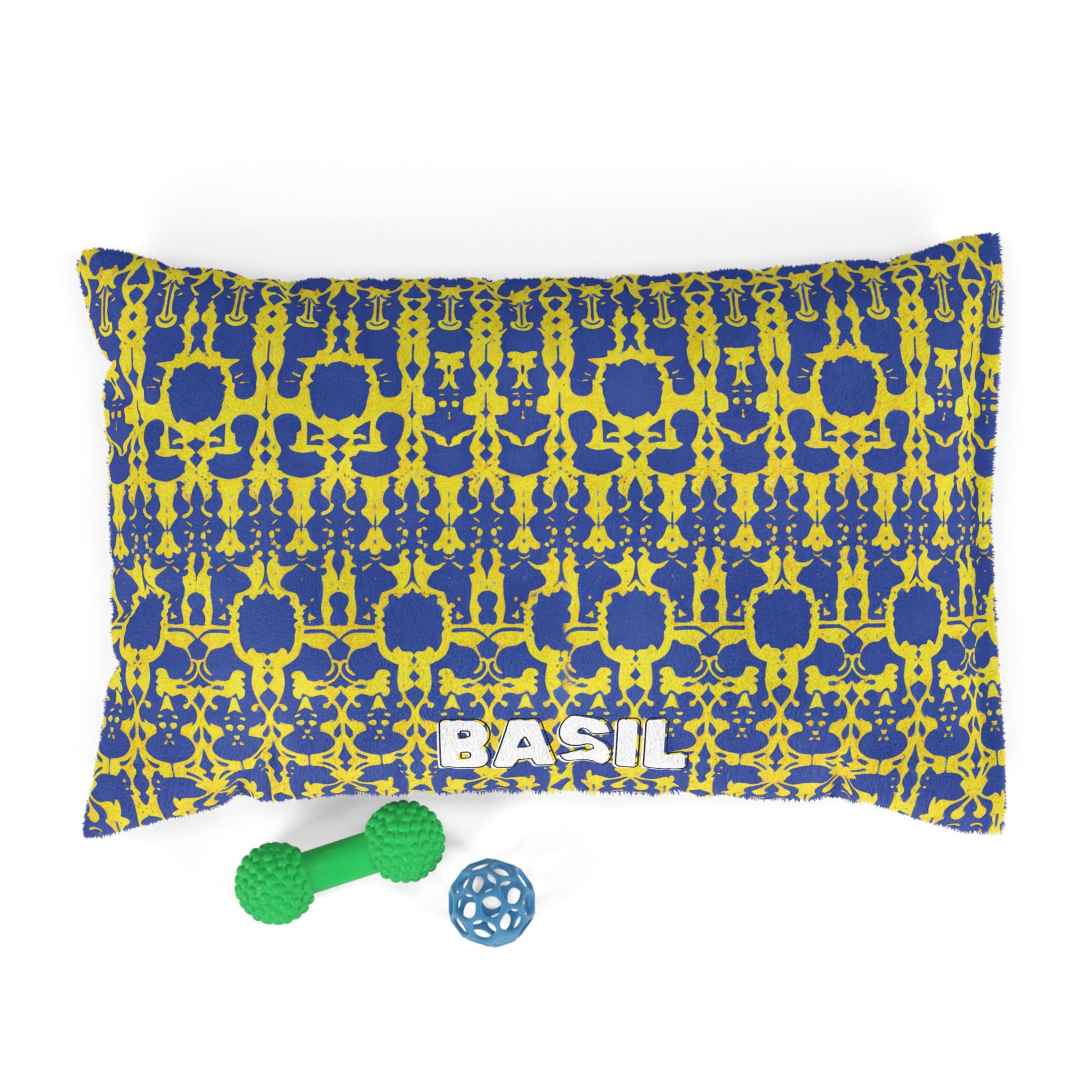 A rectangular dog bed with a yellow and blue abstract pattern and the word BASIL printed in white near the bottom showcases luxurious comfort. In front are a green bone-shaped chew toy and a small blue ball toy, all set against a white background.