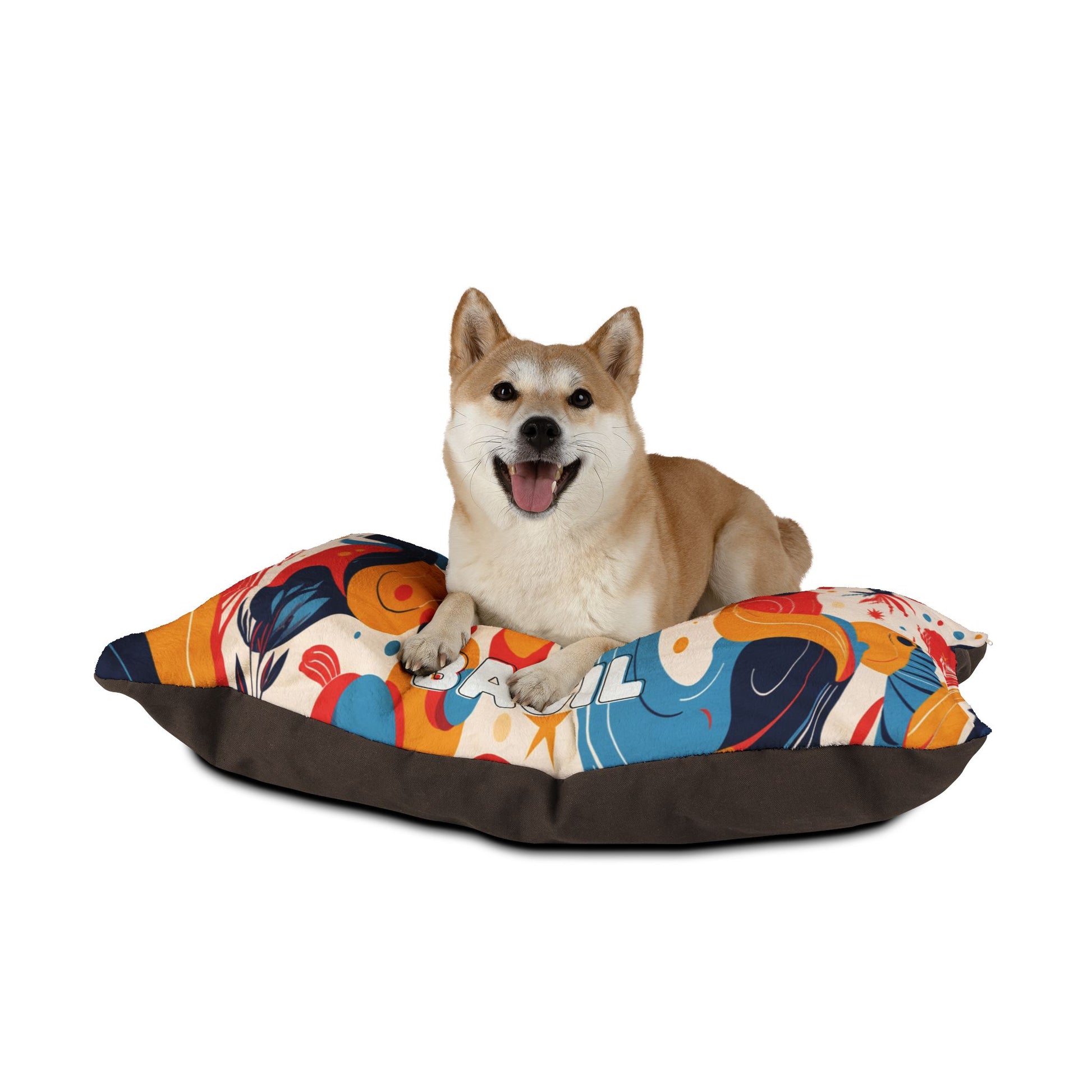 A Shiba Inu dog with tan fur lies on a colorful, African-inspired abstract-patterned pillow with dark brown sides. The dog is facing forward with mouth open and tongue out, appearing happy, against a plain white background.