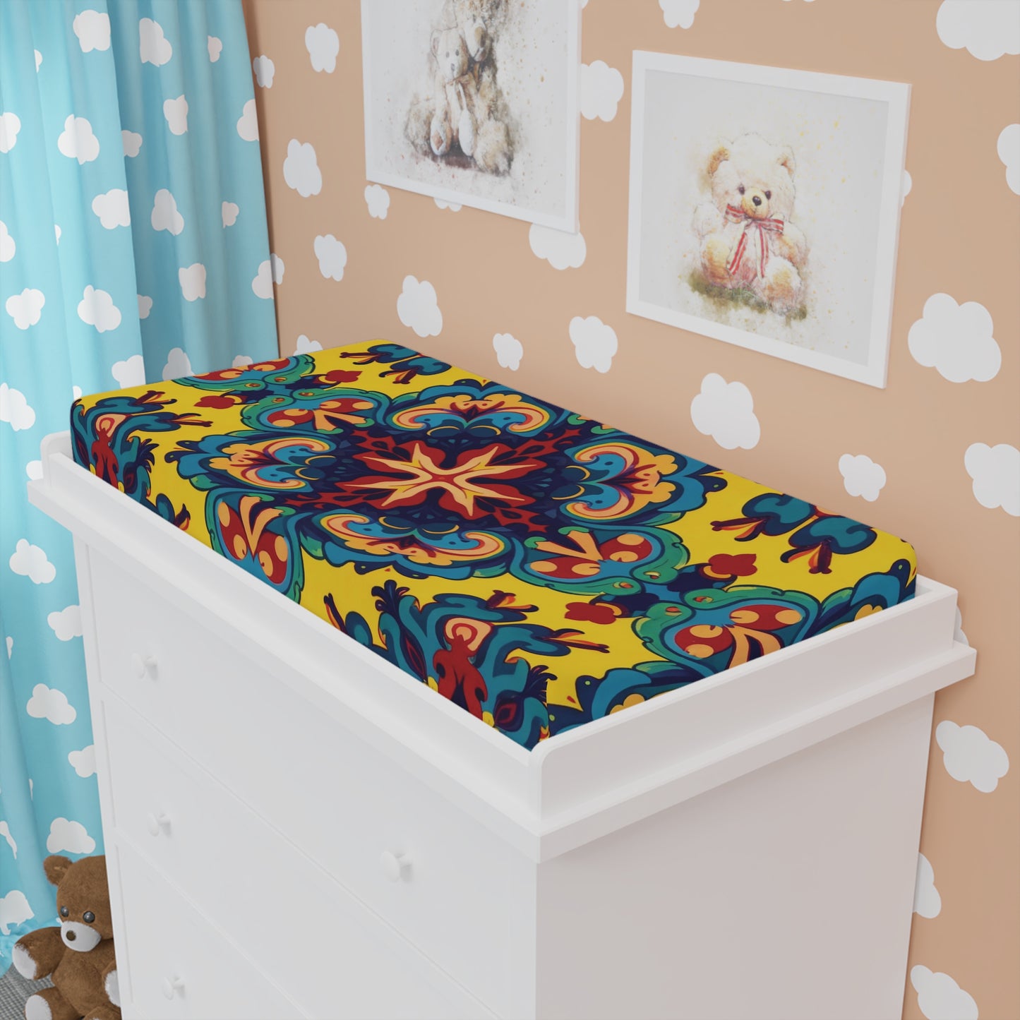 A white dresser holds a vibrant, African-inspired custom changing pad cover with ornate, symmetrical patterns in yellow, red, blue, and green. Behind it: teddy bear art and plush, cloud-print wallpaper, and blue curtains complete the cozy nursery.