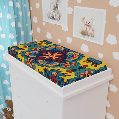 A white dresser holds a vibrant, African-inspired custom changing pad cover with ornate, symmetrical patterns in yellow, red, blue, and green. Behind it: teddy bear art and plush, cloud-print wallpaper, and blue curtains complete the cozy nursery.