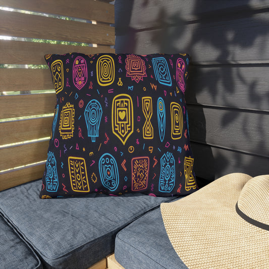 A large square pillow with colorful African-inspired abstract tribal patterns in blue, pink, and yellow on a black background sits on a gray outdoor bench, next to a beige sun hat. The setting is shaded by wooden slats and dark siding.