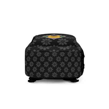 A square black ottoman with gray floral patterns and a colorful, round emblem featuring a yellow star on top, reminiscent of a stylish travel bag. The ottoman has short black feet, soft cushioning, and the floral motif wraps around its sides.