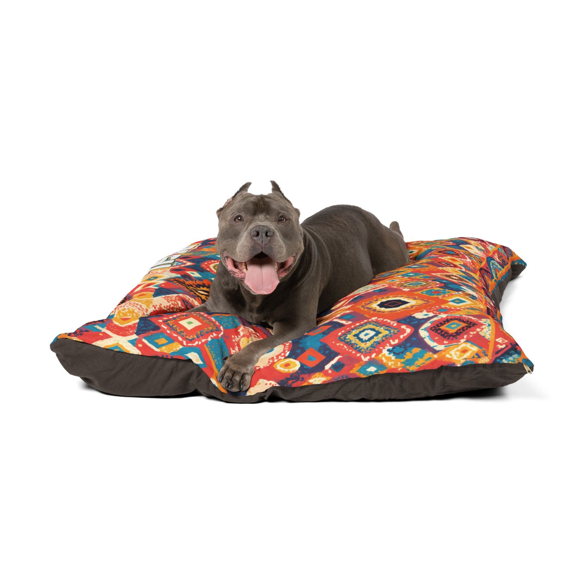 A happy grey pit bull dog with cropped ears lies on a large, African-inspired dog bed featuring red, blue, yellow, and orange geometric designs. The dog’s mouth is open with its tongue out, appearing relaxed against a white background.
