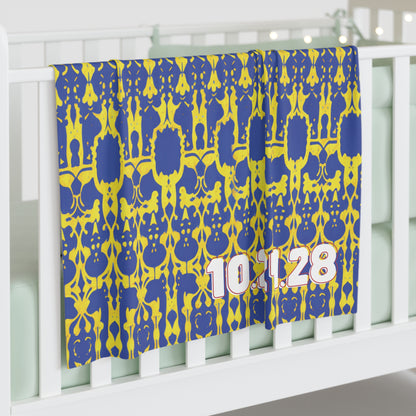 A blue and yellow patterned personalized baby blanket with the numbers 10.1.28 in large, white and pink font drapes over a white crib in a softly lit room, featuring pale bedding and decorative string lights in the background.