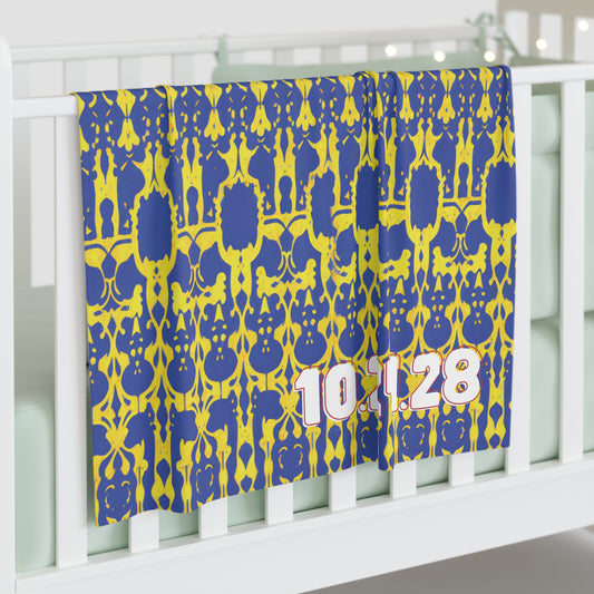 A blue and yellow patterned personalized baby blanket with the numbers 10.1.28 in large, white and pink font drapes over a white crib in a softly lit room, featuring pale bedding and decorative string lights in the background.