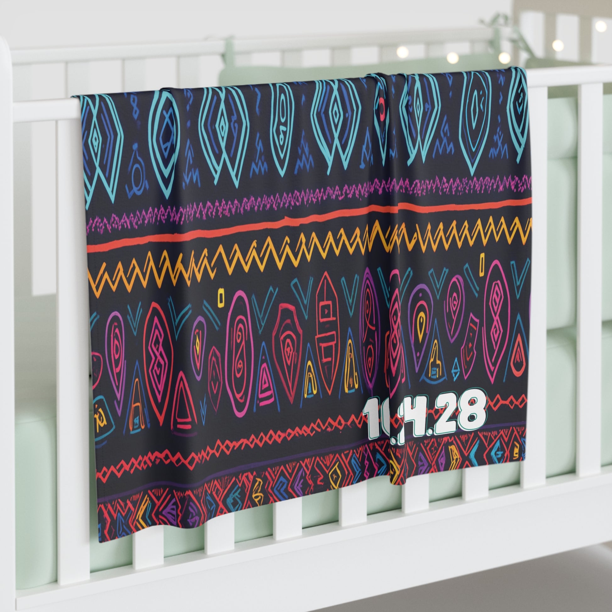A soft personalized baby blanket with colorful, African-Inspired decor patterns in blue, purple, yellow, red, and orange drapes over a white crib rail. The numbers 11.4.28 in bold white text are printed in the lower right corner of the blanket.