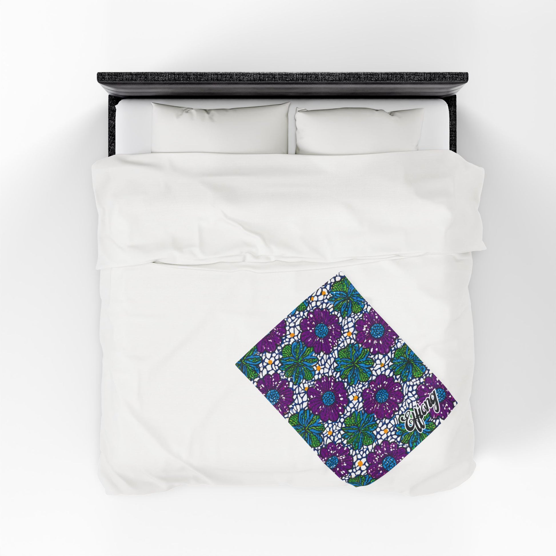 A neatly made bed with white bedding and two pillows, viewed from above. An African-inspired personalized blanket, adorned with blue, green, and purple flowers, is folded near the bottom right corner—making a unique gift for any occasion.