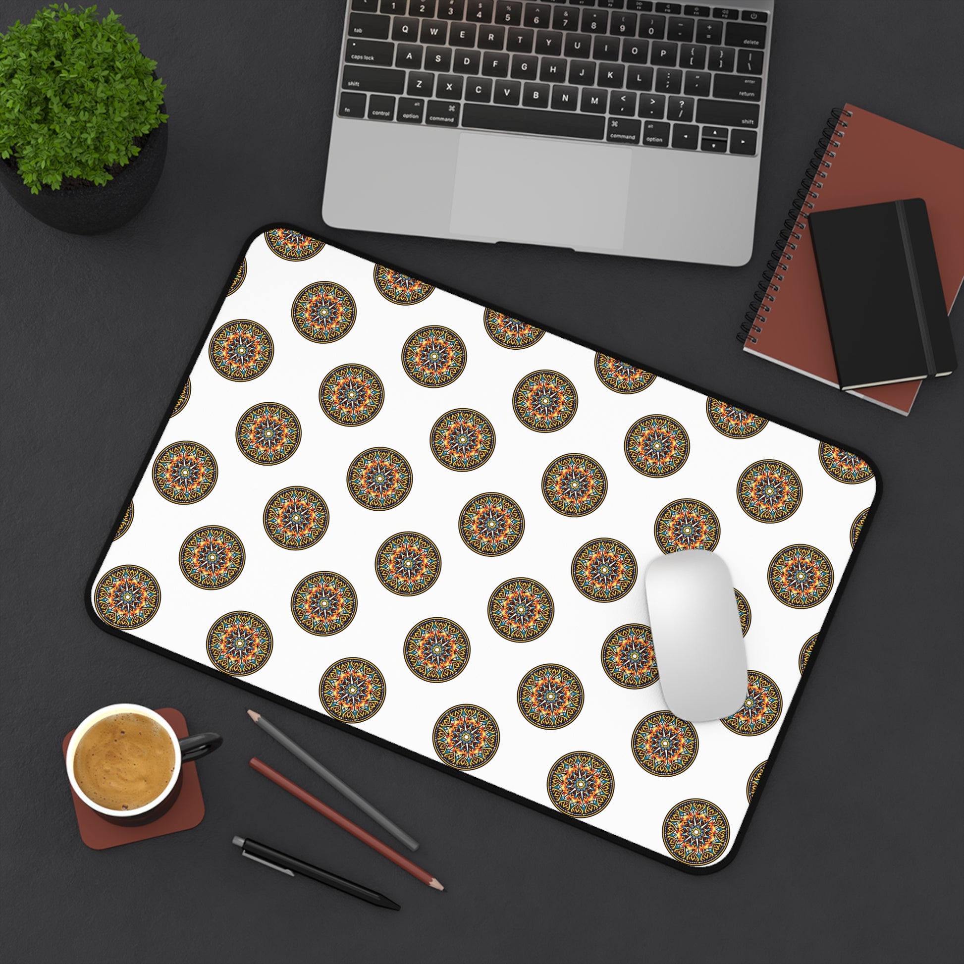 A neatly arranged black desk with a laptop, small potted plant, notebooks, pencils, coffee, and a white mouse on an ergonomic neoprene desk mat featuring a repeating colorful mandala pattern—a vibrant workspace accessory.
