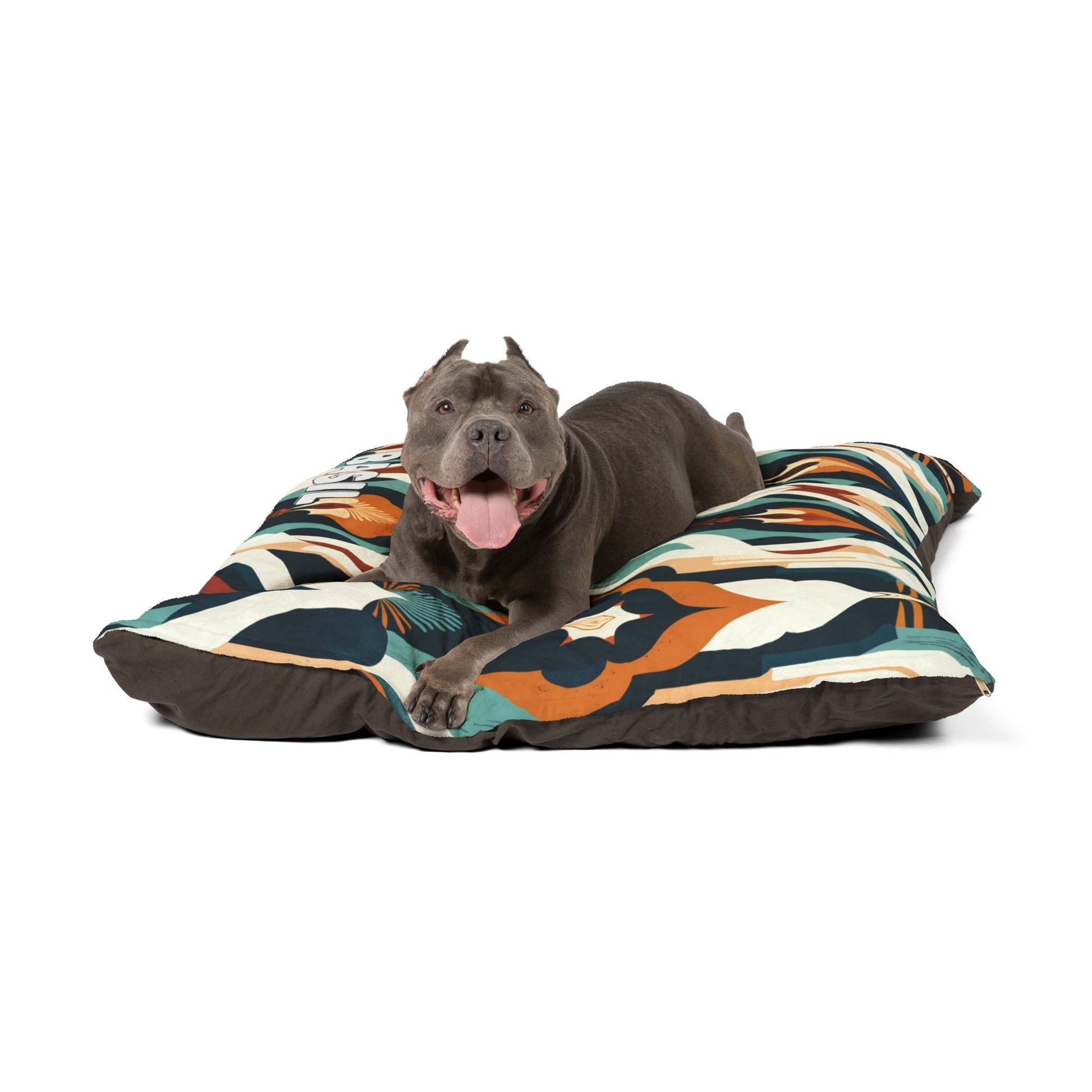 A gray pit bull with cropped ears lies comfortably on a cozy pet bed with colorful geometric patterns. The dog faces forward, mouth open and tongue out, looking happy and relaxed against a white background.
