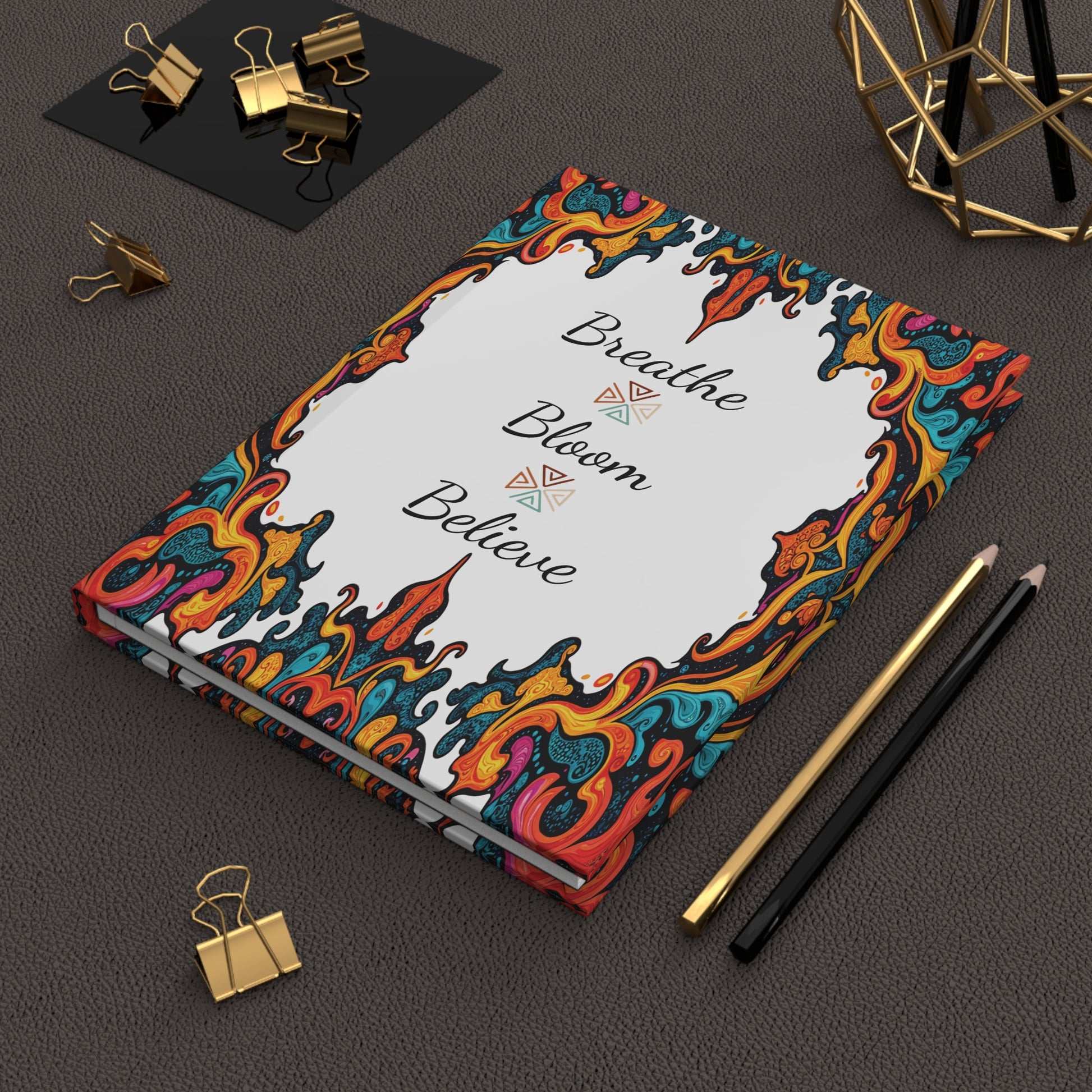 A colorful, ornate African-inspired journal with Breathe Bloom Believe on the cover sits on a textured dark surface. Nearby are gold binder clips, black and gold pencils, and a geometric decor piece, all neatly arranged.