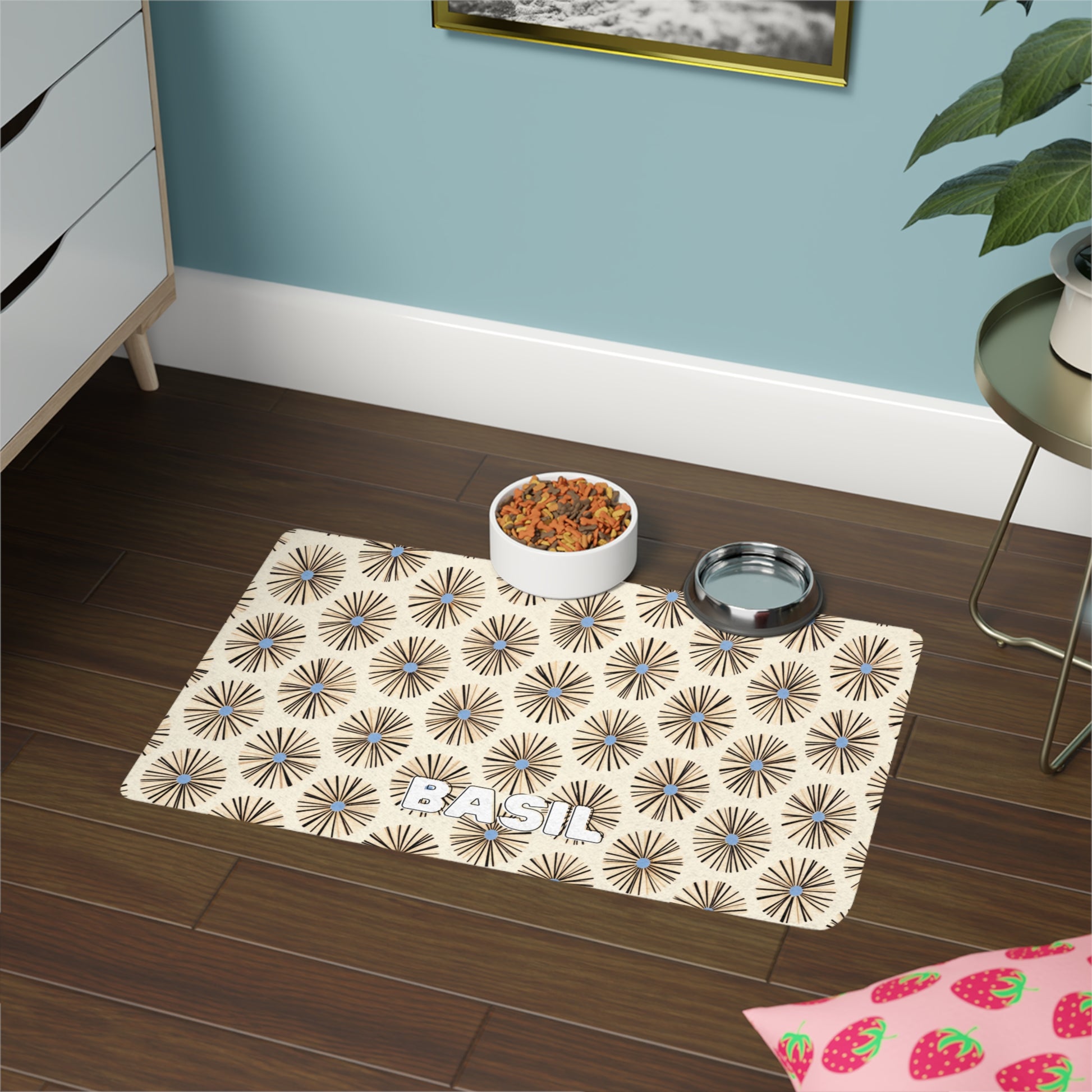 A beige pet food mat with a black, African-inspired starburst pattern and the name “BASIL” lies on a wood floor. On the mat are a white bowl of pet food and a silver water bowl, near a plant, strawberry-patterned item, and dresser against a teal wall.