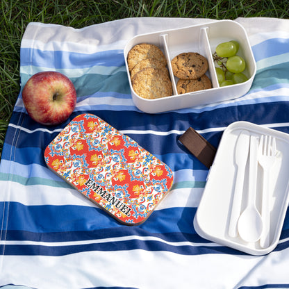 A personalized African-inspired bento lunch box with cookies and green grapes sits on a blue striped picnic blanket beside an apple. The lid, decorated with vibrant floral patterns and the name Emmanuel, rests near a white cutlery case on the grass.