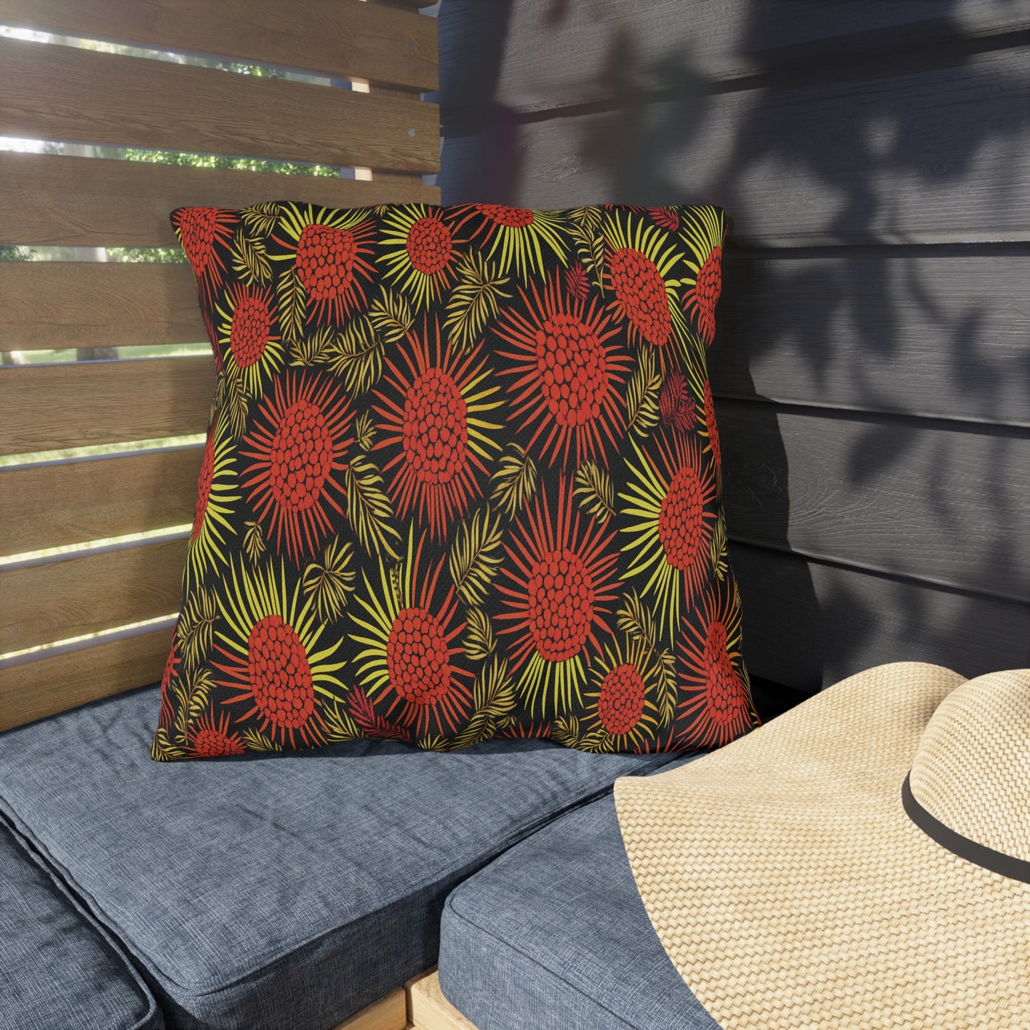 A square pillow with an African-inspired bold red and yellow floral pattern on a black background rests on a blue cushioned bench. Nearby, a straw hat lies on the seat as sunlight and shadows fall across wooden slats and a dark wall behind.