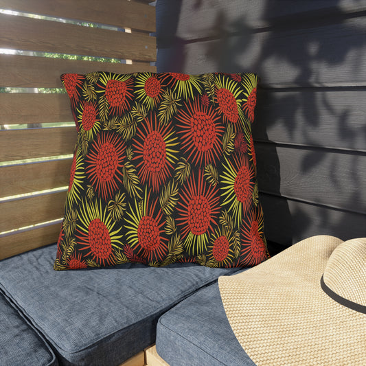 A square pillow with an African-inspired bold red and yellow floral pattern on a black background rests on a blue cushioned bench. Nearby, a straw hat lies on the seat as sunlight and shadows fall across wooden slats and a dark wall behind.