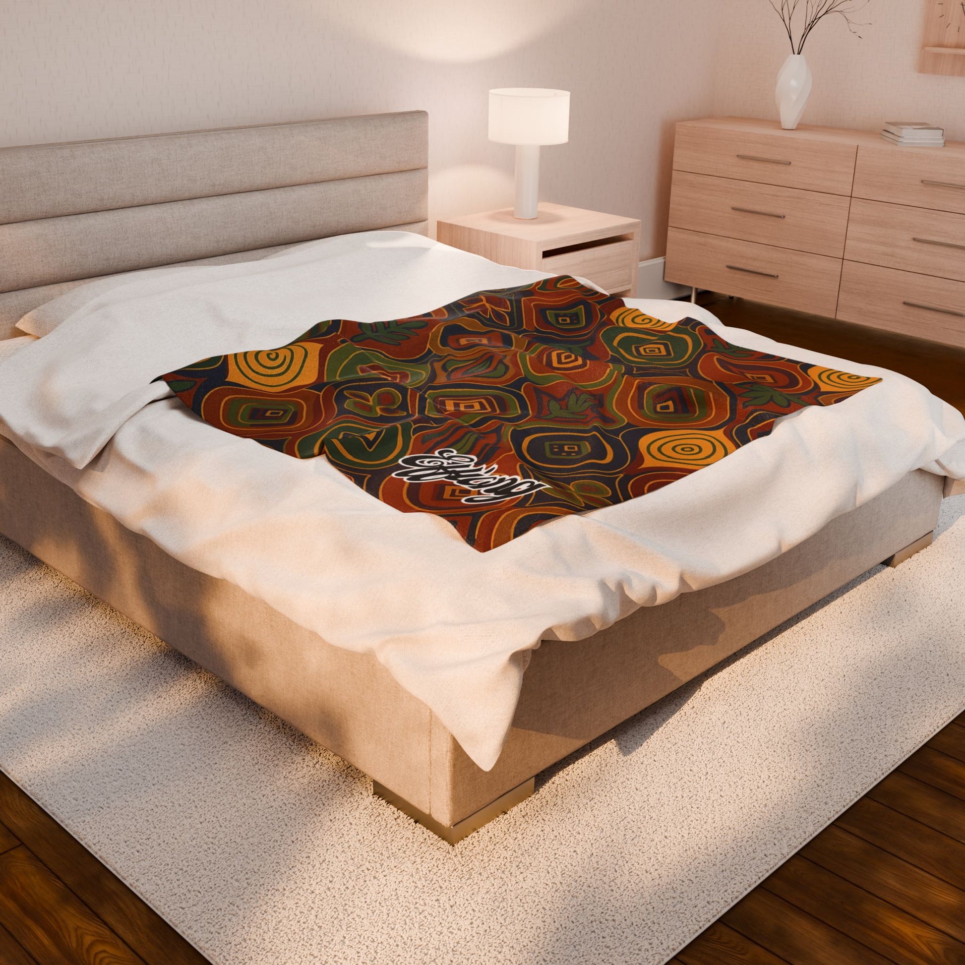 A neatly made bed in a modern, softly lit bedroom features an African-inspired personalized blanket with colorful swirls and geometric shapes laid across the foot. The word “Ebony” appears on this unique keepsake gift, complementing the light wood furniture and white rug.