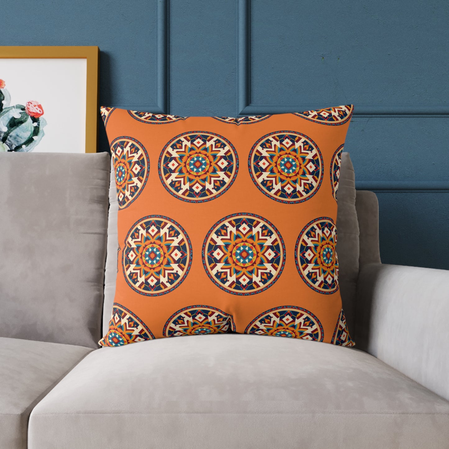 A vibrant home decor piece, this large throw pillow showcases African-inspired mandala patterns in blue, gold, white, and black on vivid orange fabric. It sits on a light gray sofa with a deep blue wall and framed cactus art in the background.