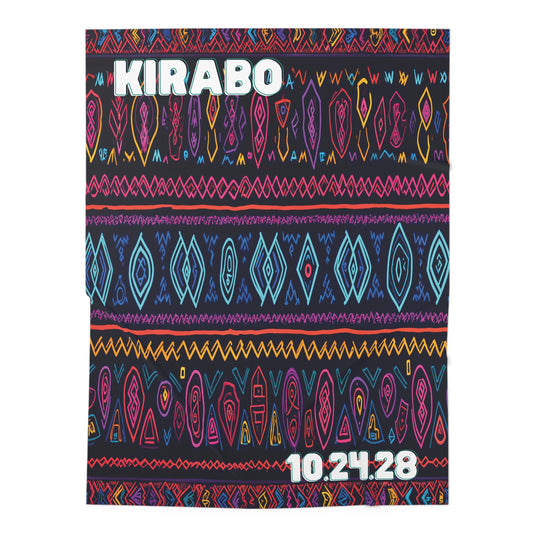 A personalized baby blanket features KIRABO in large white letters at the top and 10.24.28 in smaller numbers at the bottom right, with colorful, African-inspired geometric patterns in blue, pink, yellow, and orange on a dark base.