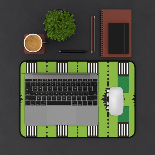 A top-down view of a workspace with a laptop and white mouse on a green patterned neoprene desk mat, a coffee cup, small potted plant, two pencils, pen, and stacked brown notebooks with a black stripe, all on a dark surface.