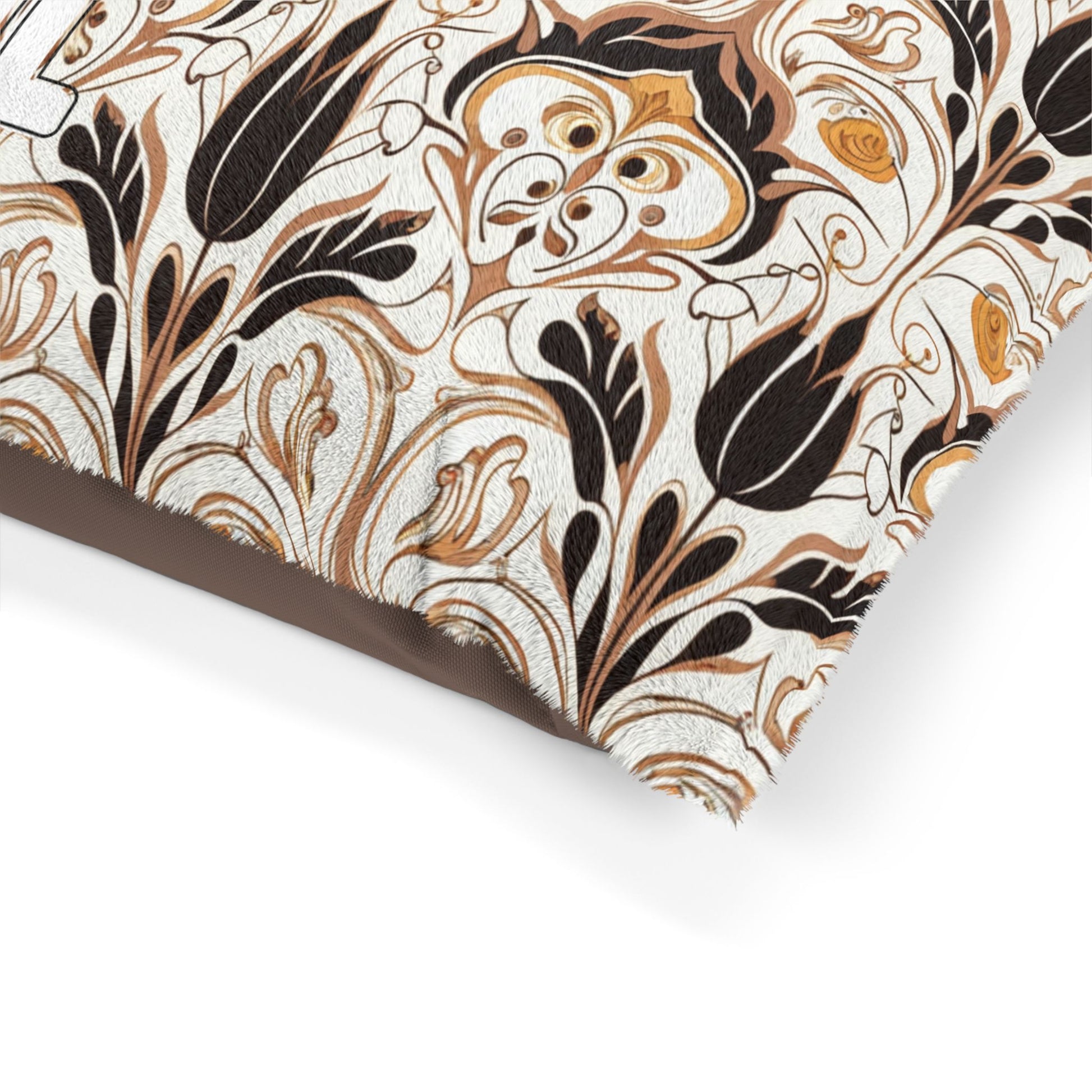 A close-up of a decorative pillow corner with a fringed edge. The fabric features an African-inspired, intricate floral and leaf pattern in shades of black, brown, and beige on a cream background. Part of a bold white letter is visible on the surface.