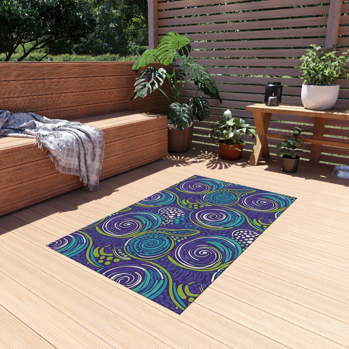 A vibrant outdoor patio features a modern wooden bench with a gray plaid blanket, several potted plants, and a colorful weather-resistant rug in purple, blue, and green on light wood flooring. Sunlight streams through slatted wood fencing.