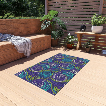 A vibrant outdoor patio features a modern wooden bench with a gray plaid blanket, several potted plants, and a colorful weather-resistant rug in purple, blue, and green on light wood flooring. Sunlight streams through slatted wood fencing.