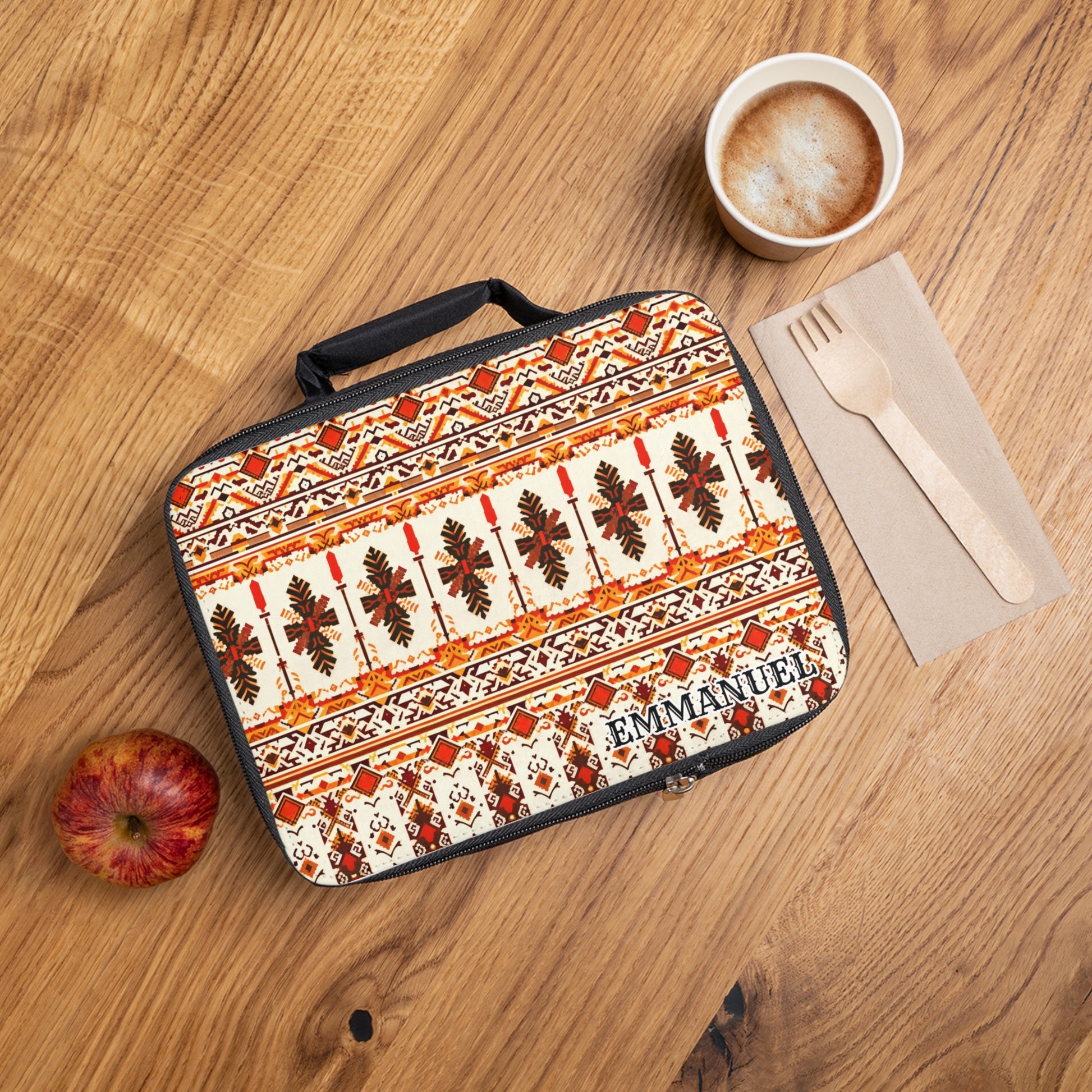 A patterned, insulated lunch tote labeled “EMMANUEL” sits on a wooden table next to a red apple, foam-topped coffee, and wooden cutlery on a brown napkin. The lunch bag features an African-inspired orange and brown geometric design.