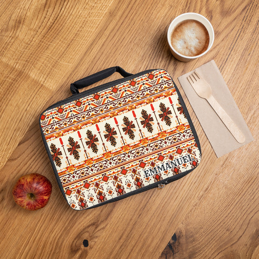 A patterned, insulated lunch tote labeled “EMMANUEL” sits on a wooden table next to a red apple, foam-topped coffee, and wooden cutlery on a brown napkin. The lunch bag features an African-inspired orange and brown geometric design.