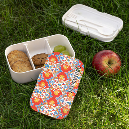 A colorful, African-inspired lunch box with a red, floral-patterned lid labeled EMMANUELLE sits open on grass. Inside, three sections hold cookies, a chocolate chip cookie, and green grapes. Nearby are a red apple and BPA-free container with cutlery.